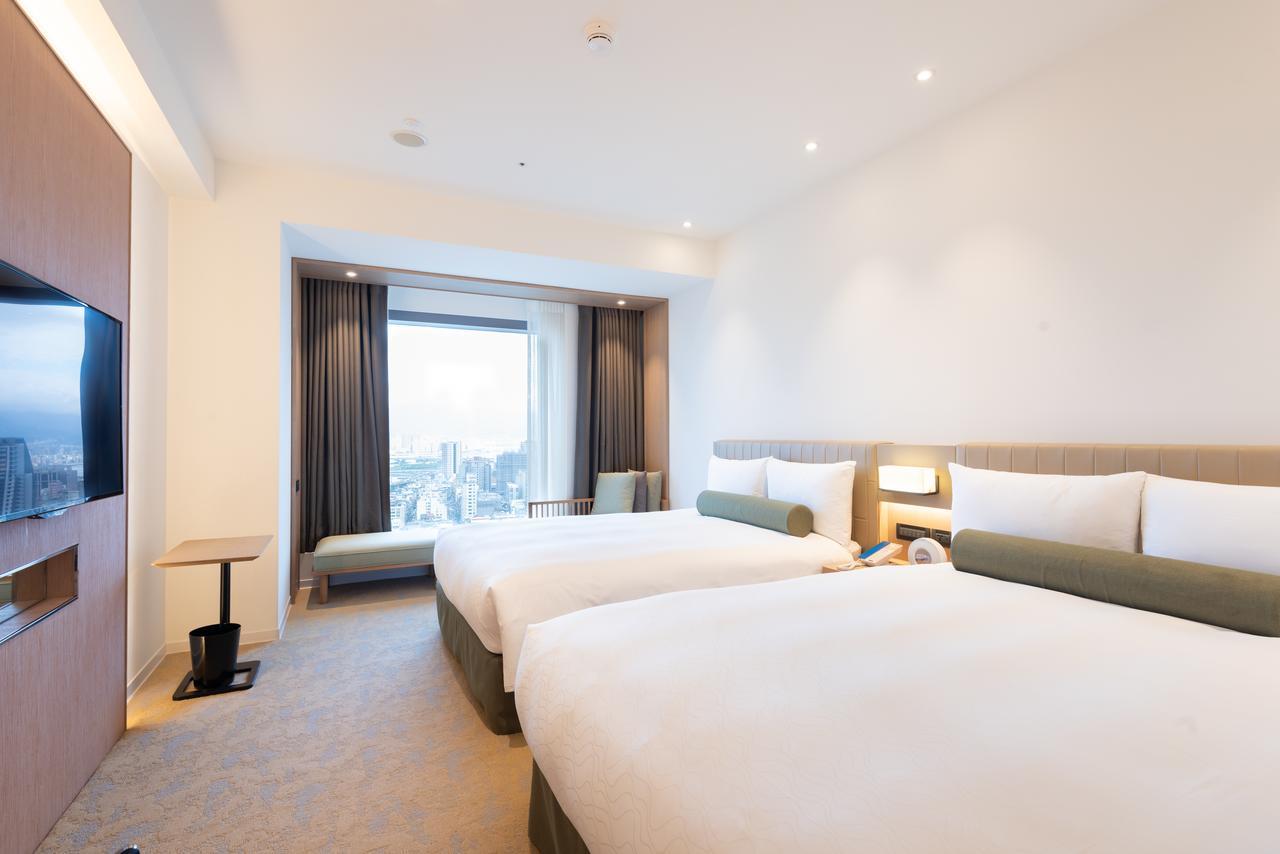 Caesar Metro Taipei Hotel Luaran gambar The photo shows a modern hotel room featuring two twin beds with white bedding and green accent pillows. The walls are painted in light colors, creating a spacious and bright atmosphere. A large window allows natural light to enter, offering a view o