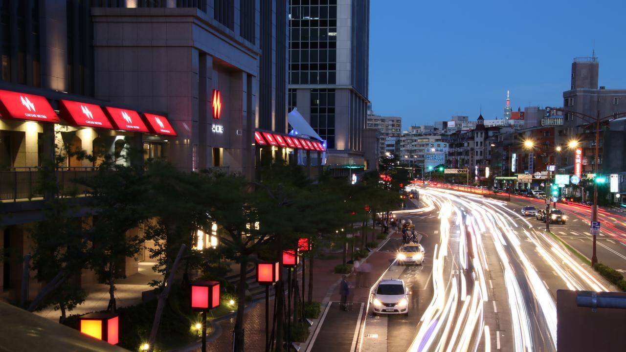 Caesar Metro Taipei Hotel Luaran gambar The photo shows a city scene during twilight. In the foreground, there is a busy road with streaks of light from moving vehicles, suggesting it's a lively area. On the sides of the street, red lanterns or light fixtures are visible, adding to the amb
