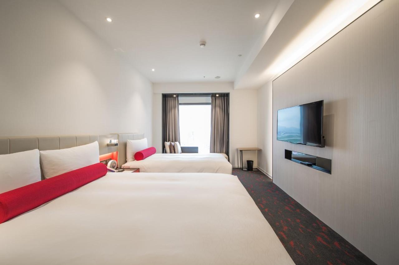 Caesar Metro Taipei Hotel Luaran gambar The photo shows a modern hotel room featuring two large beds with white linens and red accent pillows. There is a window with curtains that allow natural light to enter the room. On the wall opposite the beds, there is a flat-screen television mounte